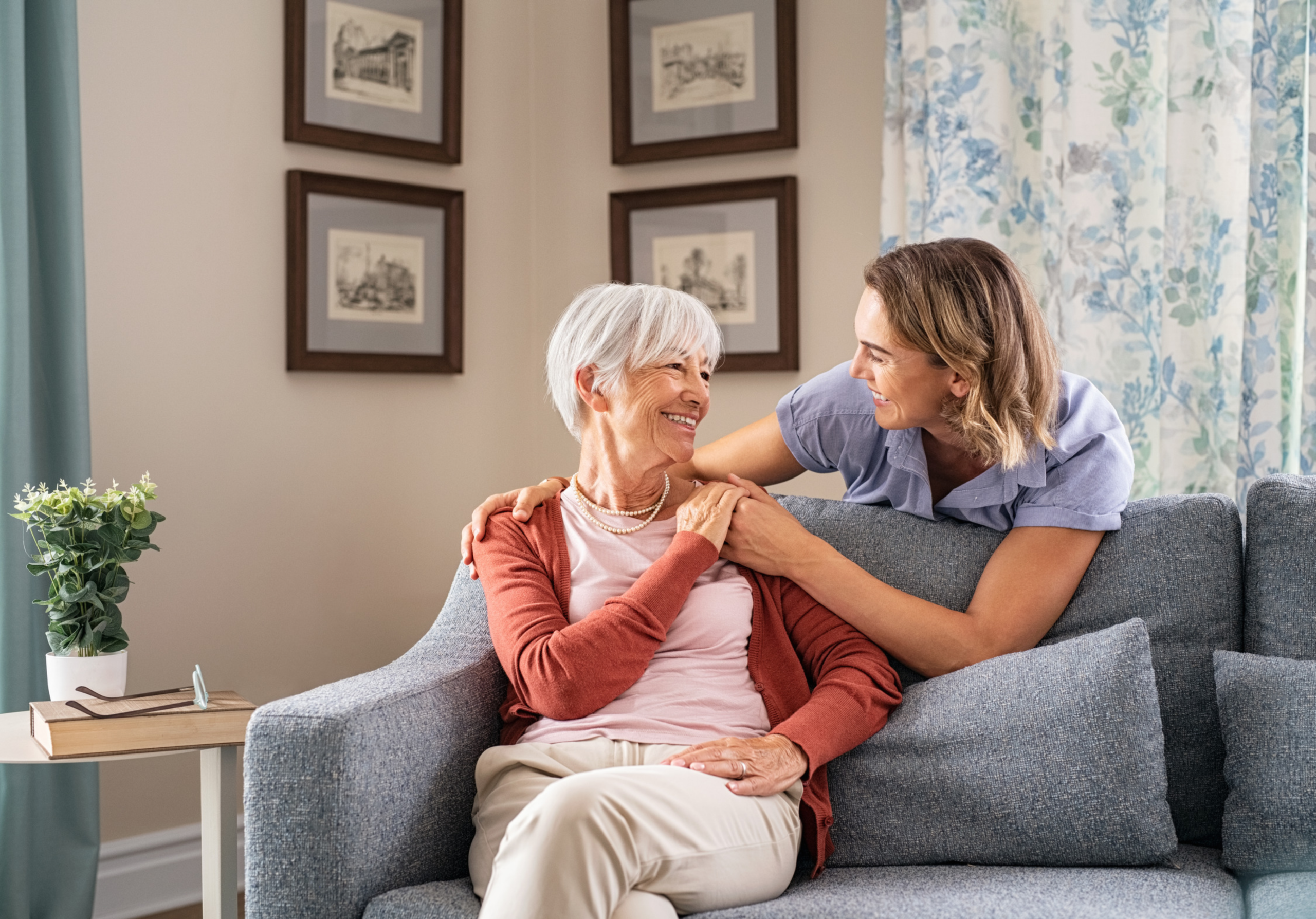 An elderly lady sitting on the sofa being comforted by her smiling daughter.