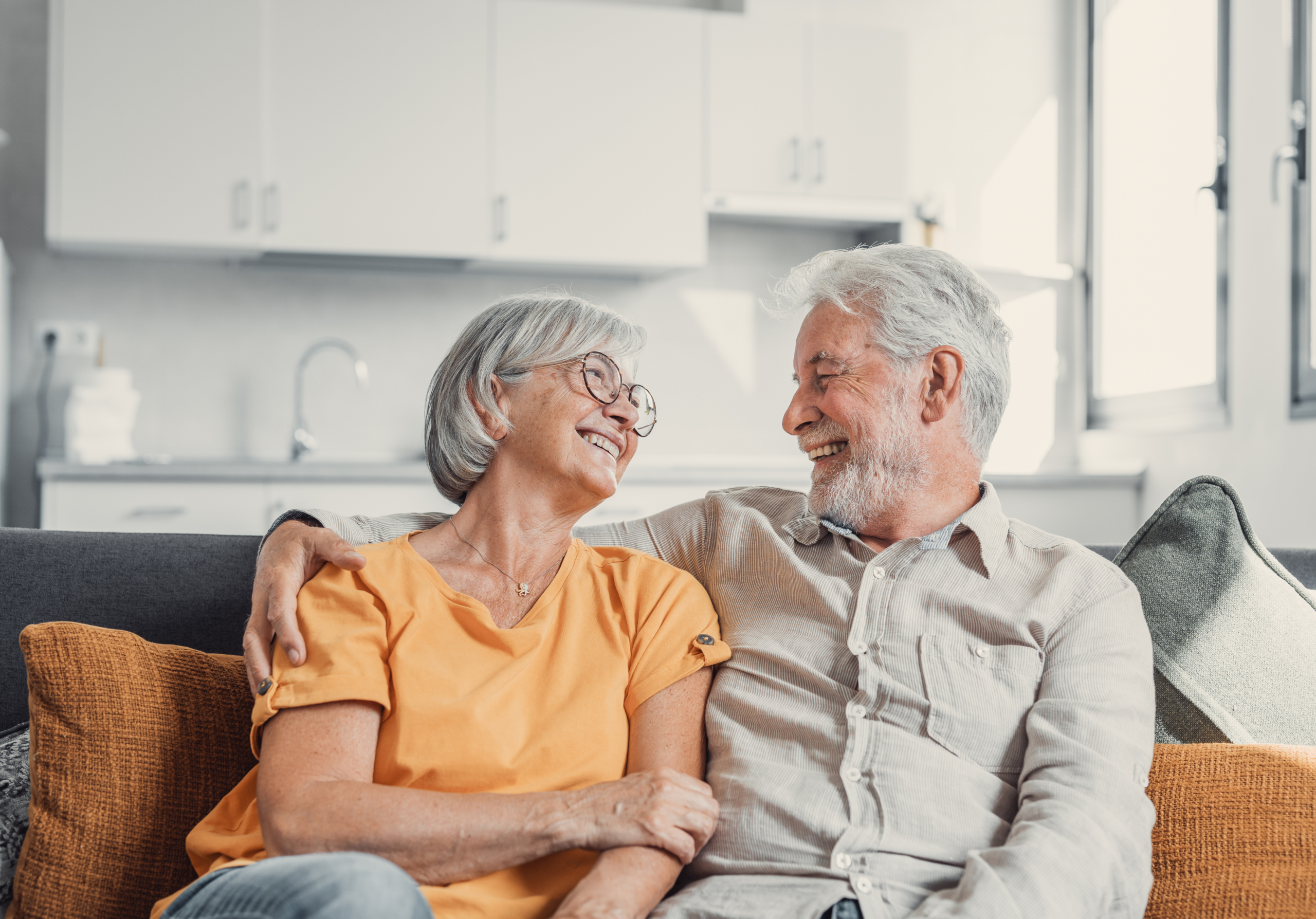 Older couple sitting on a sofa, smiling and gazing into one another's eyes.